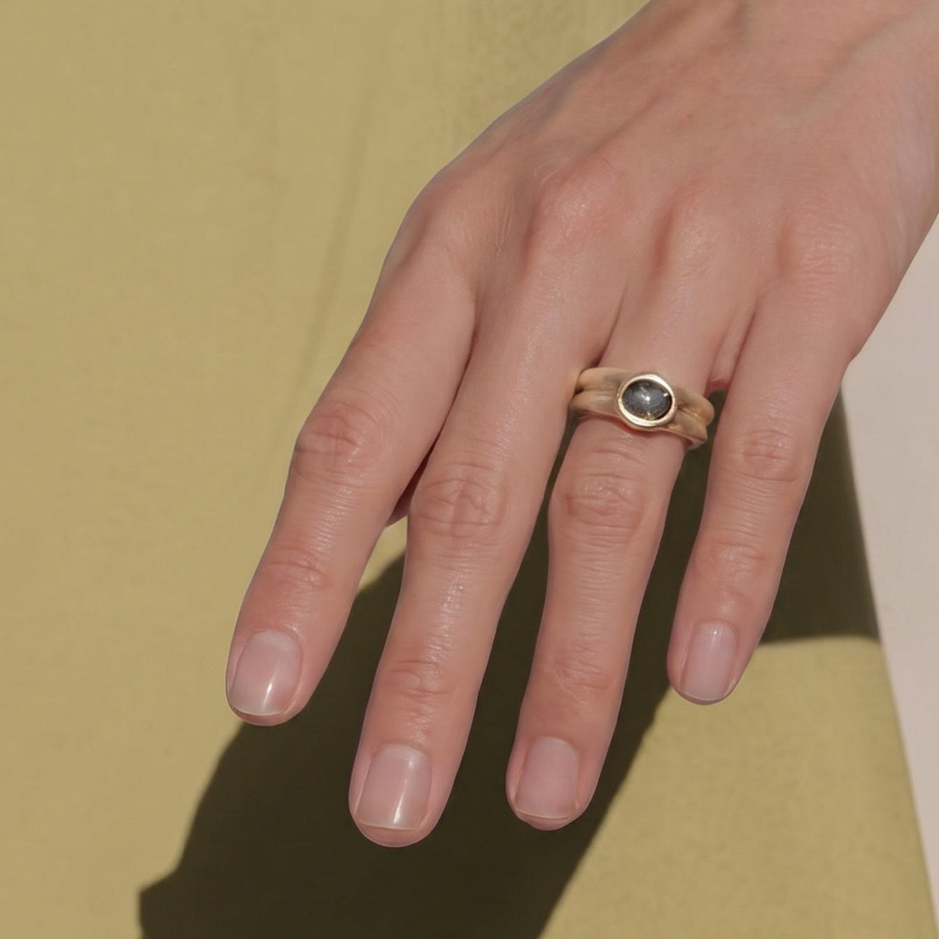 A close-up of a person's hands resting on a yellow surface, with a gold ring featuring a round dark green stone worn on one finger. Strong shadows of the hands are cast on the surface below.